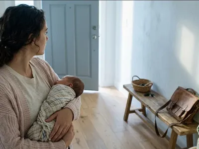 A person sitting on a bed with shoulders slightly dropped, a soft blanket nearby, in quiet exhaustion, representing the themes of "Career Burnout in New Mothers: Does It Get Better?".