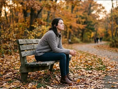 A woman sitting alone on a park bench surrounded by autumn leaves, gazing into the distance with a pensive expression, evoking the open-ended and nonlinear nature of grief.