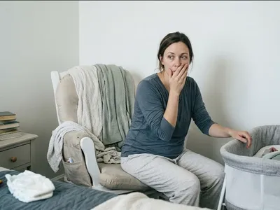 A mother sitting on the edge of a bed in morning light, infant asleep nearby in a bassinet, representing the themes of "How to Talk to Someone About Your Postpartum OCD".