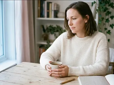 A person at a table with a warm drink, looking forward with a calm, open expression, representing the themes of "Recovering from Birth Trauma: What Healing Actually Involves".