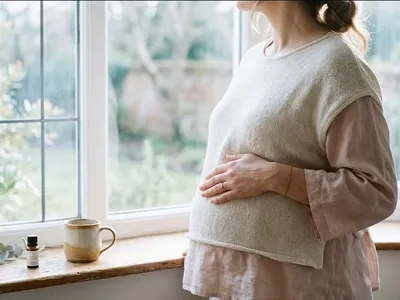 A woman standing near a window, one hand on her belly, soft backlight, representing the themes of "TFMR Grief Support: Navigating the Unique Heartbreak of Ending a Wanted Pregnancy".