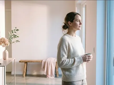 A figure seated on a window seat, looking out at a grey sky, a soft blanket nearby, representing the themes of "What to Say to Someone Who Had a Miscarriage: Words That Help".